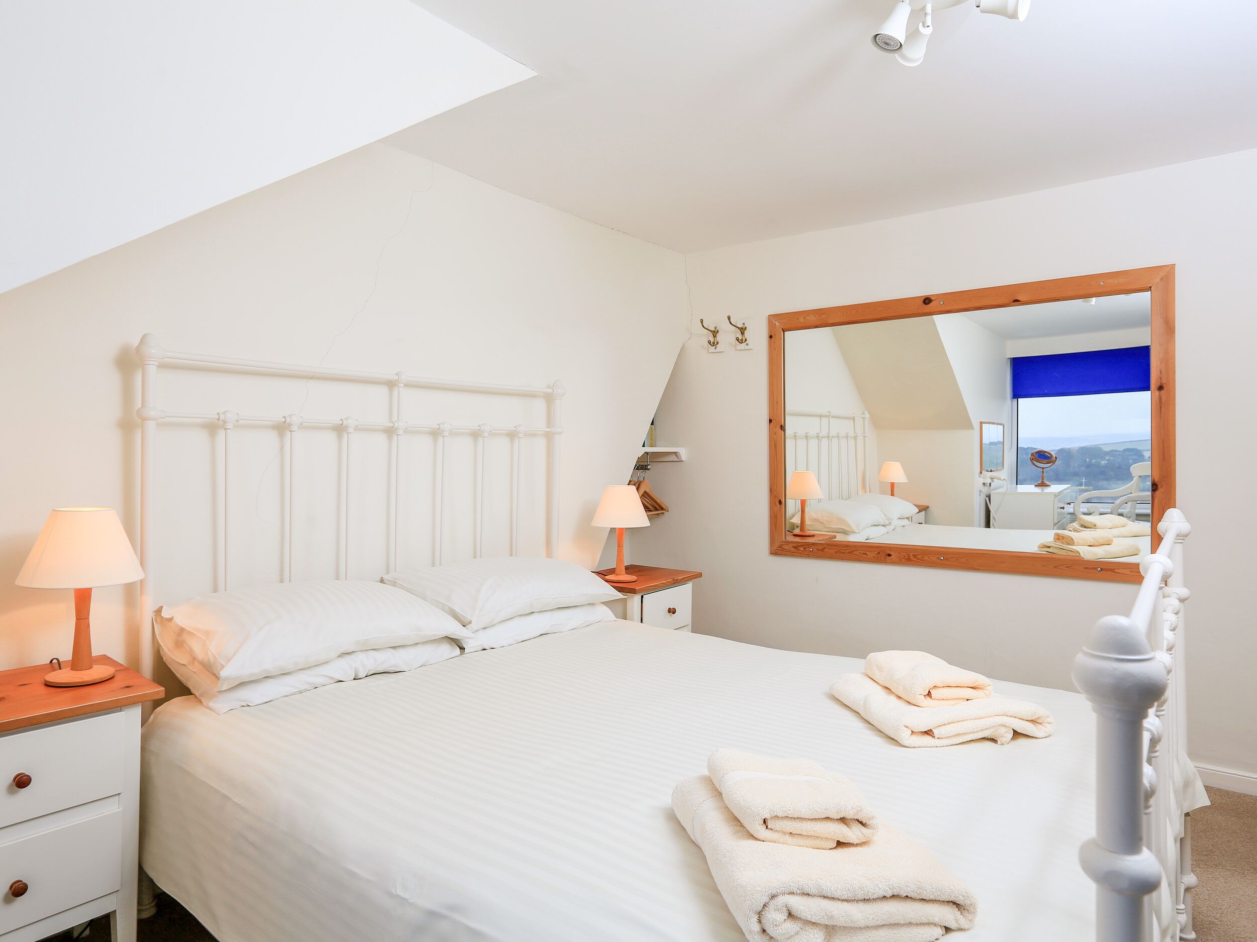 Une chambre avec un lit blanc, deux tables de chevet, un grand miroir et une vue sur l’extérieur.