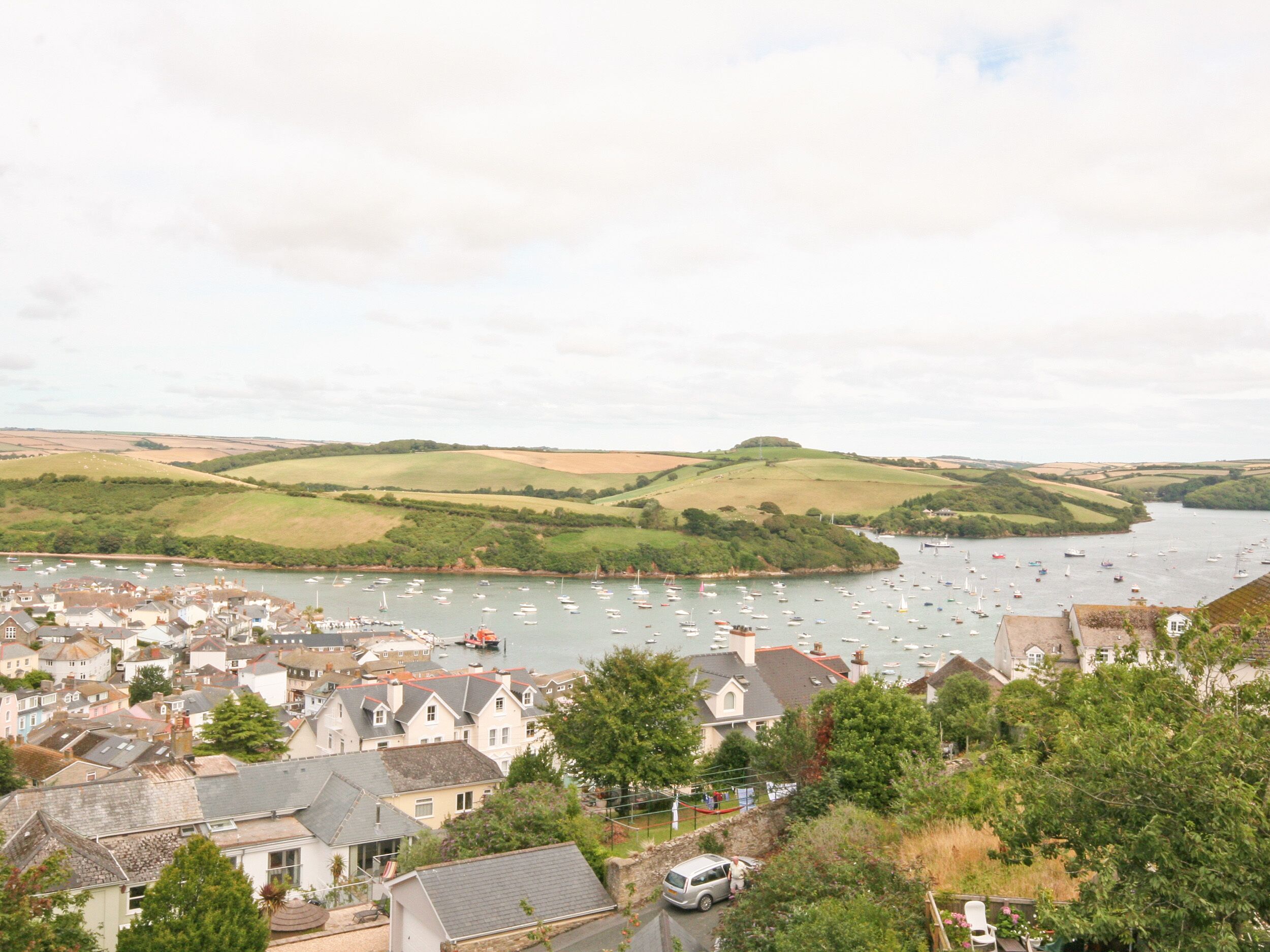 A coastal town with a marina filled with boats, surrounded by rolling hills and houses with traditional architecture.