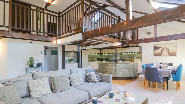 A spacious living area with a grey sofa, a wooden coffee table, and a dining table set for four. A kitchen is visible in the background. - Barley Mews (Weymouth)