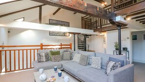A spacious living room with a grey sofa, wooden beams, a staircase, and a dining area with a table set for tea. - Barley Mews (Weymouth)