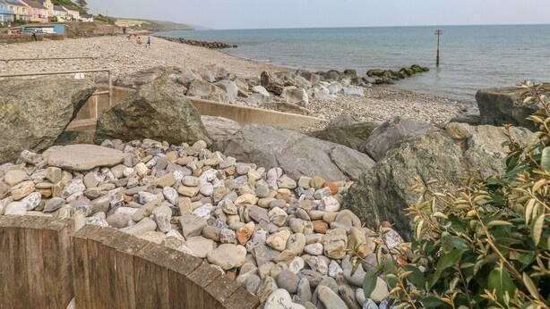 Una spiaggia rocciosa con una passerella in legno che conduce al mare, case sullo sfondo e un cielo sereno.