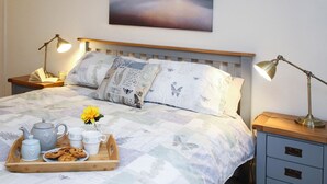 A neatly made bed with a floral duvet, two bedside lamps, a tray with a teapot, cups, and pastries, and a painting of a landscape above the bed. - Gwyndy Bach (Caernarfon)