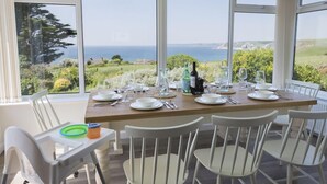 A dining area with a table set for six, large windows offering a view of the sea and greenery, and two pendant lights above the table. - Seaspray (Kingsbridge)
