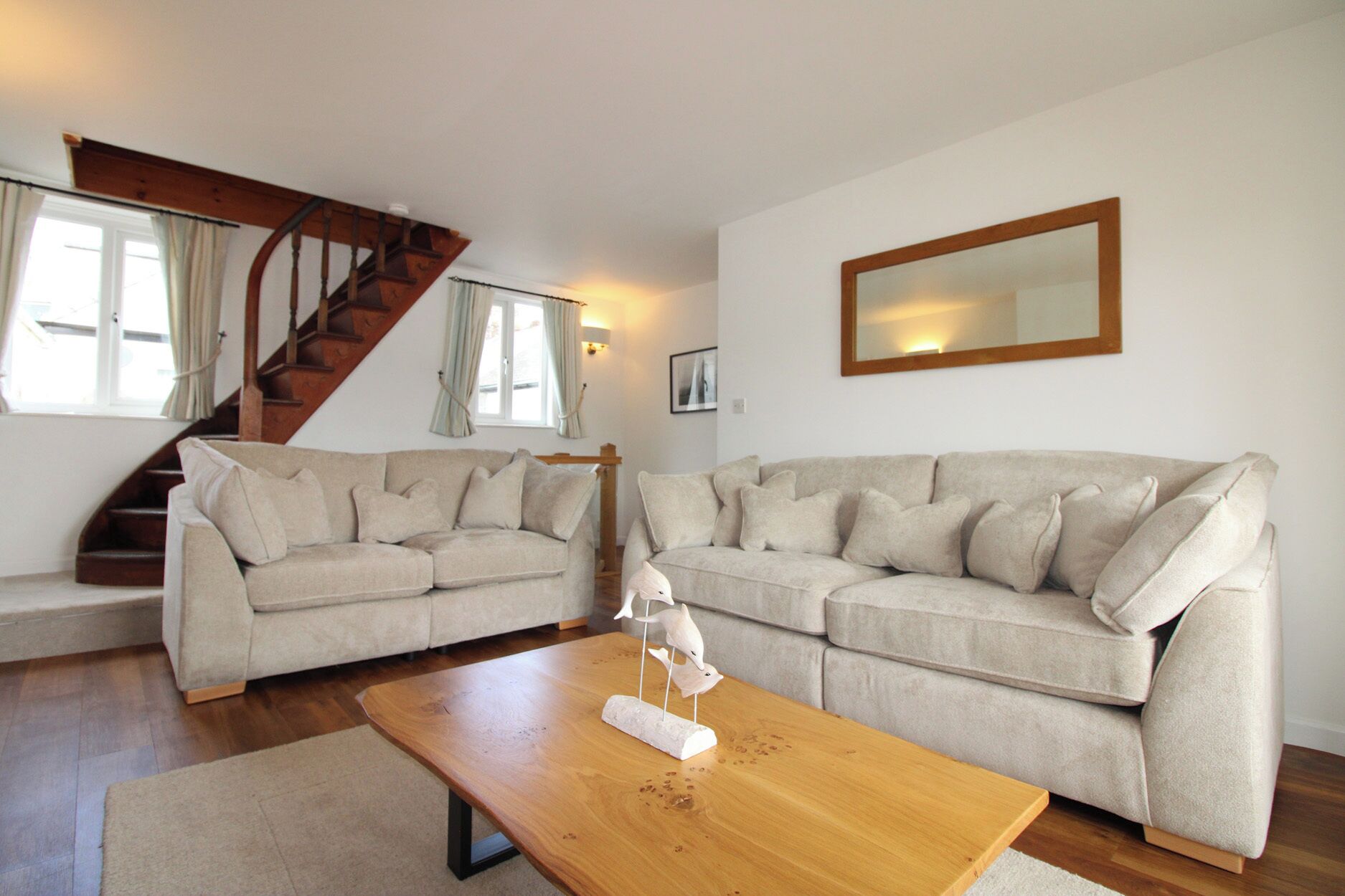 Een woonkamer met een houten trap, twee beige banken, een houten salontafel en een spiegel aan de muur.