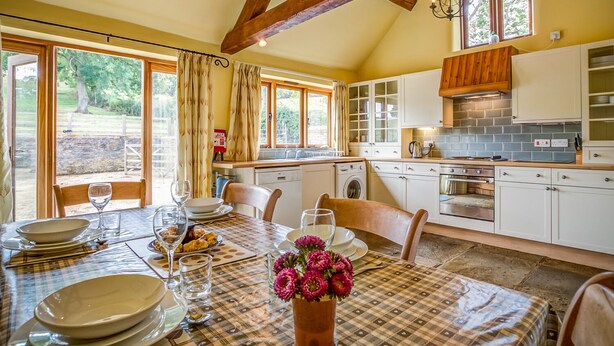Un coin repas avec une table dressée pour quatre personnes, une cuisine équipée d’appareils modernes et de grandes fenêtres offrant une vue sur l’extérieur.