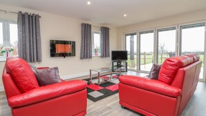A modern living room with two red leather sofas, a flat-screen TV, and a large window offering a view of the outdoors. - The Donkey Shed (Sherborne)