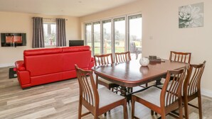 A modern living room with a red sofa, wooden dining table, and chairs, large windows with curtains, and a flat-screen TV mounted on the wall. - The Donkey Shed (Sherborne)