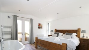 A bedroom with a wooden bed, a white bathtub, and a wooden nightstand with a lamp. - Beech Tree Lodge (York)