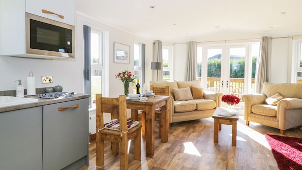 A modern kitchen with a built-in oven and microwave, wooden dining table with chairs, a sofa, and a red rug. - Beech Tree Lodge (York)