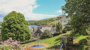 A playground with slides and climbing structures, surrounded by a wooden fence and lush greenery. - Westland (Salcombe)