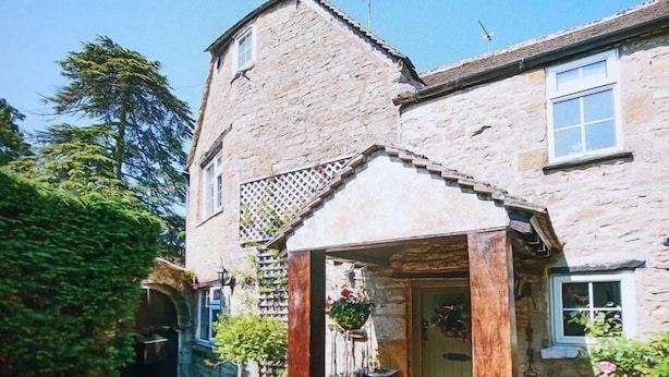 A stone house with a wooden porch, a small garden, and a clear blue sky. - Pike Cottage (Cheltenham)