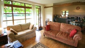 Una sala de estar con un sofá color beige y otro sofá estampado, piso de madera y una cocina al fondo.