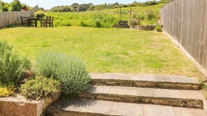 A grassy backyard with a wooden table and chairs, stone steps leading up to a gate, and a wooden fence. - Studland Cottage (Swanage)