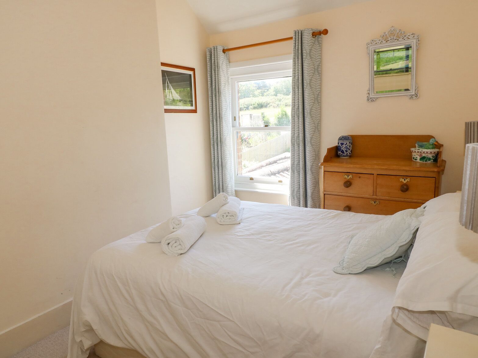 Una cama bien tendida con sábanas blancas y toallas enrolladas, un aparador de madera con adornos, y una ventana con cortinas.