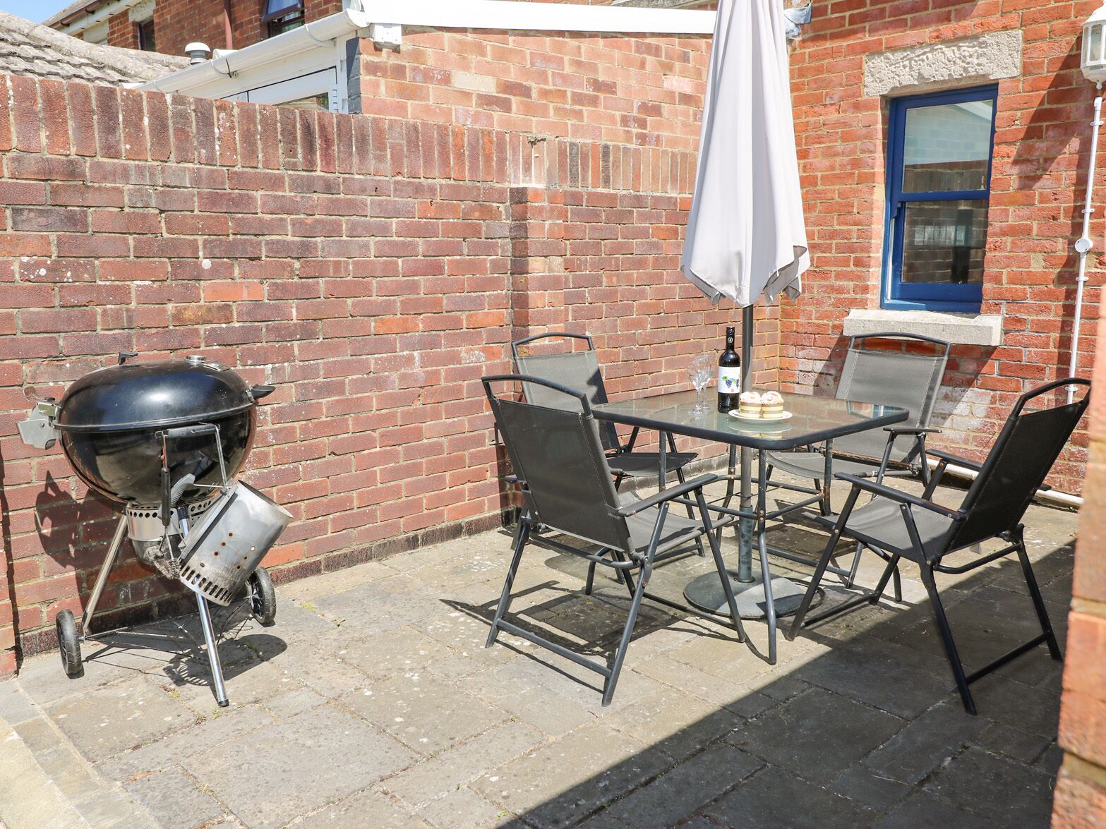 Un patio con una parrilla, mesa exterior, sillas y una ventana con marco azul.