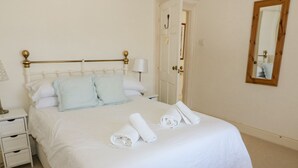 A neatly made bed with white linens, a bedside table with a lamp, and a mirror on the wall. - Studland Cottage (Swanage)