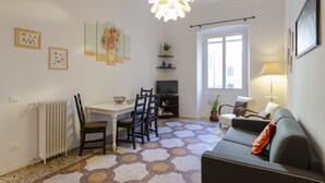 Ett vardagsrum med en soffa, stolar, ett matbord och en tv. Det finns konstverk på väggarna och en taklampa.