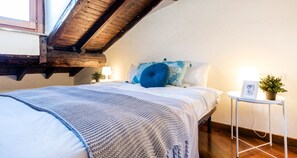 A bedroom with a sloped ceiling, a bed with a blue and white blanket, a small bedside table with a plant, and a framed picture.