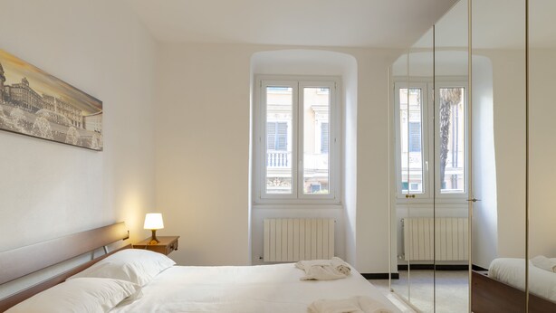 A hotel room with a large bed, a bedside table with a lamp, a window with shutters, and a mirror on the wall. - JOIVY Monte Suello (Genoa)
