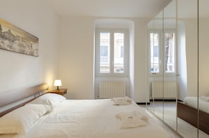 Una habitación de hotel con una cama grande, una mesita de noche con lámpara, una ventana con contraventanas y un espejo en la pared.