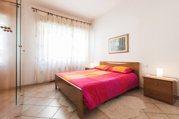 A bedroom with a bed, a nightstand, a lamp, a painting on the wall, and a glass door. - JOIVY Elly's Terrace-old City (Viareggio)