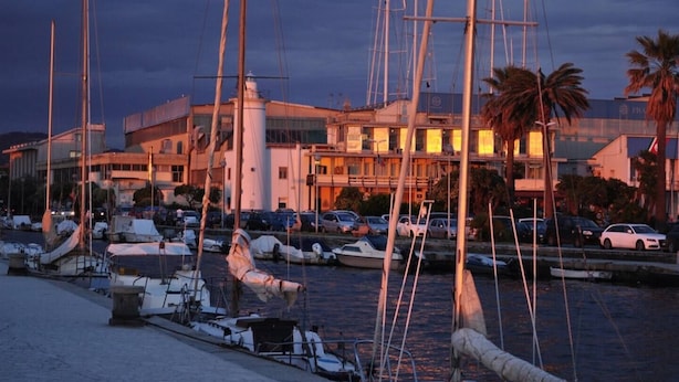Un port esportiu amb embarcacions amarrades, un edifici amb façana moderna i una palmera al primer pla.