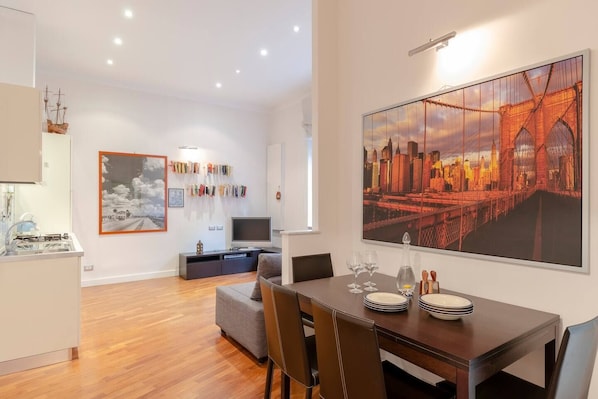 Un espace de vie moderne comprenant un coin repas, une cuisine et un salon. On y trouve des livres et un téléviseur accrochés au mur, ainsi qu’un grand tableau représentant un pont.