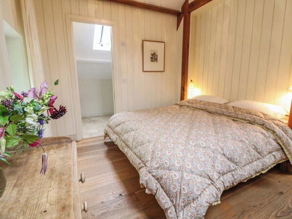 A bedroom with a wooden floor, a bed with a floral-patterned duvet, a wooden nightstand, and a vase with flowers. - The Cart Barn (Salisbury)