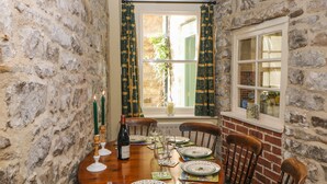 Uma sala de jantar com uma parede de pedra, uma mesa de madeira posta para quatro pessoas e uma janela com cortinas.