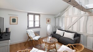 Een gezellige kamer met een donkergrijze bank, rieten stoelen, een klein rond tafeltje met een kaars, en een raam met witte gordijnen.
