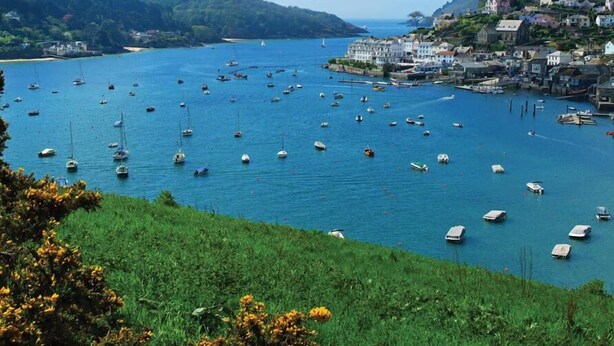 A coastal town with numerous boats anchored in the harbor, surrounded by lush greenery and hills.