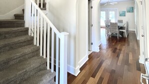 A staircase with white railings leading to a second floor, a dining area with a table and chairs, and a hallway with wooden flooring. - Gated 30A Home ☀️ 6 Lots from Pool | Easy Walk to Beach ☀️Outdoor Living Area w/ TV (Santa Rosa Beach)