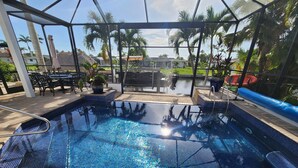 Una piscina con solárium empedrado, rodeada de palmeras y una zona de estar cubierta.