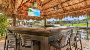 Zona de bar al aire libre con techo de paja, mostrador de madera y taburetes.