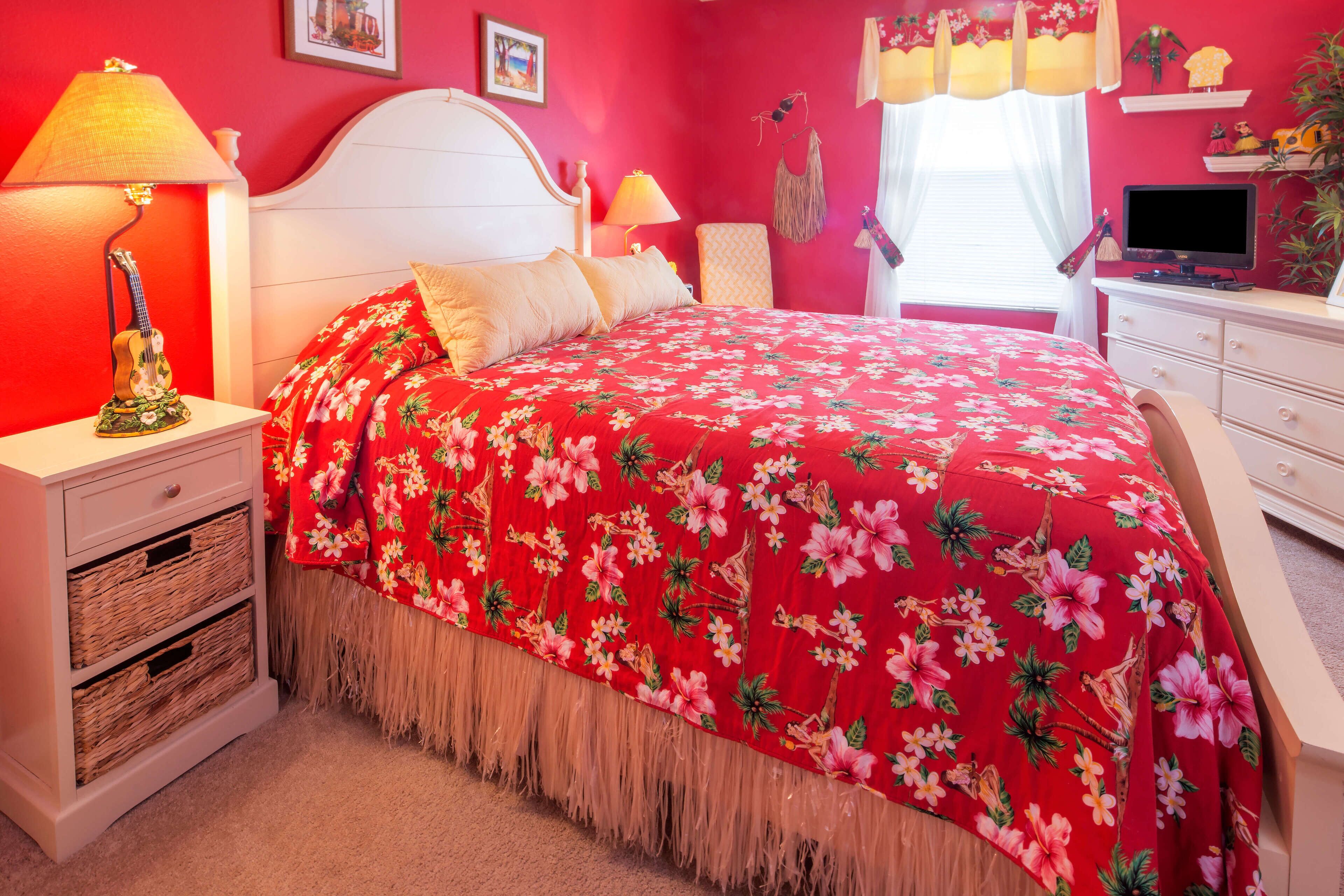 A bedroom with a red floral bedspread, white headboard, bedside lamps, and a TV on a dresser.