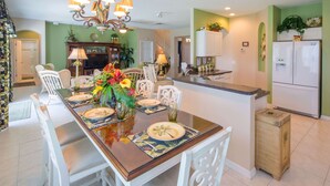 Uma sala de jantar com mesa de madeira posta para quatro pessoas, cercada por cadeiras brancas. Um lustre pendurado acima e uma televisão fixada na parede.