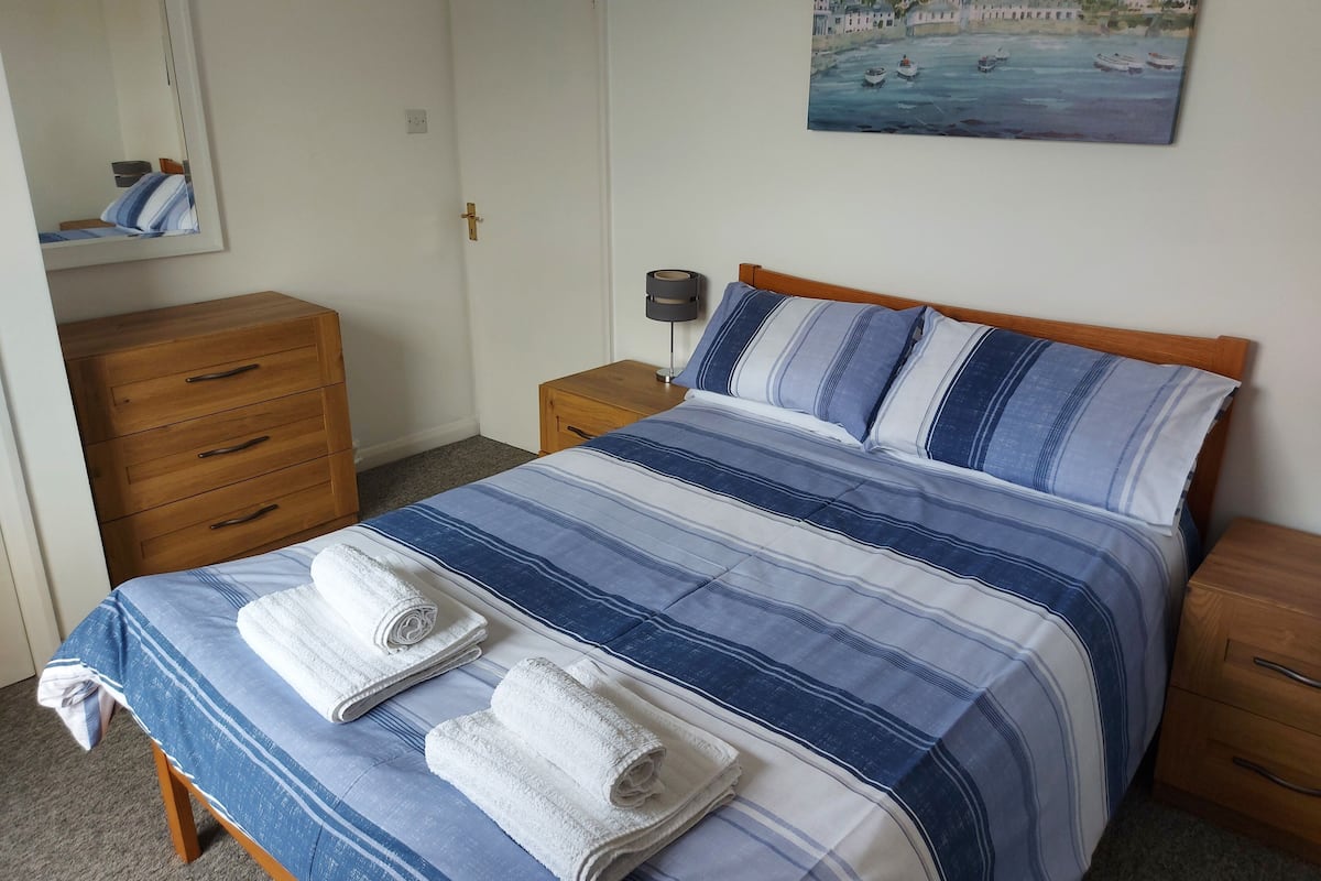 A neatly made bed with blue and white striped bedding, two rolled towels on the bed, a wooden dresser, a bedside table with a lamp, and a painting of boats on the wall.