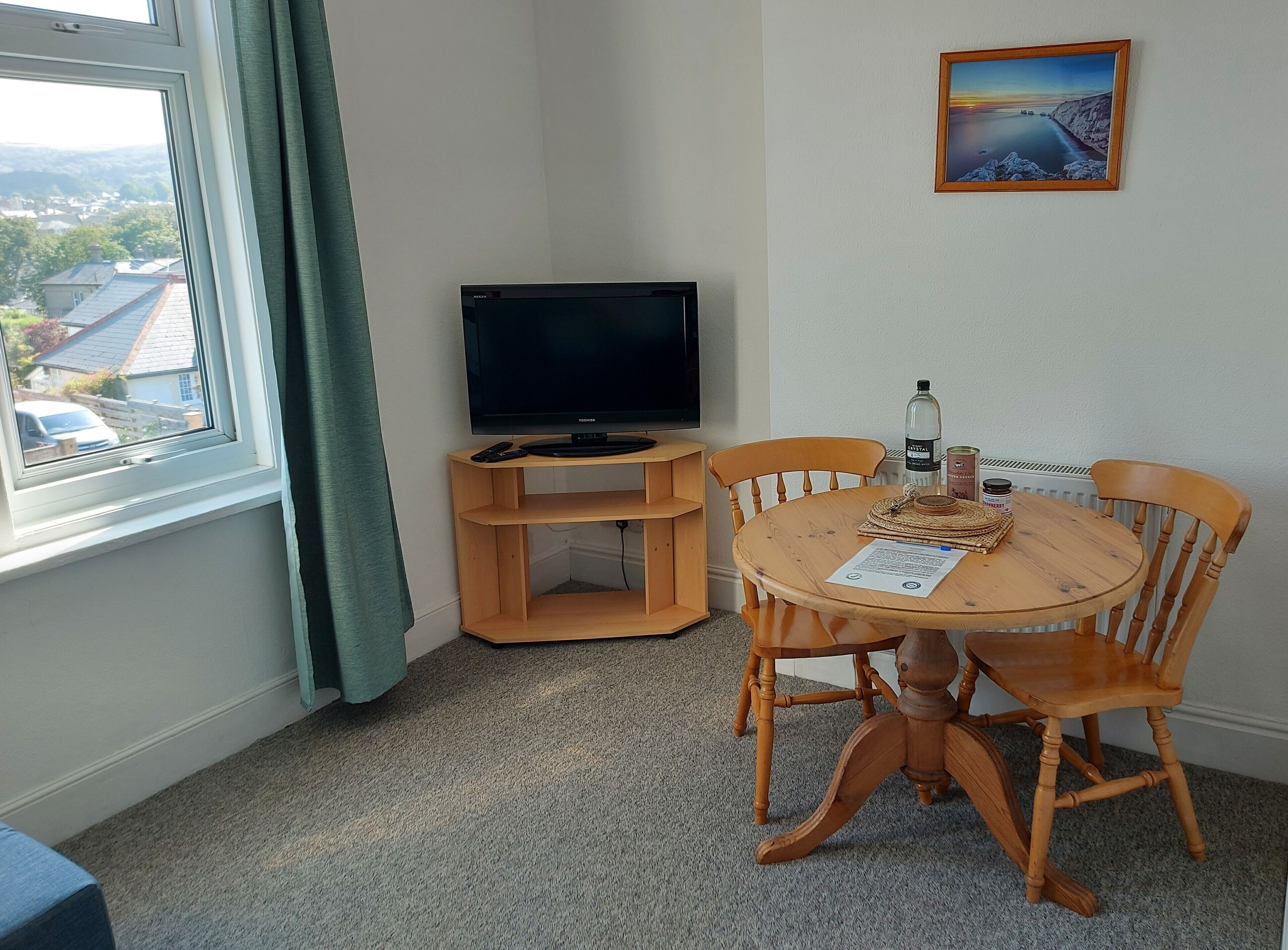 Una habitación con una televisión sobre un soporte de madera, una mesa de comedor con sillas, una ventana que da a vistas de casas y un cuadro en la pared.