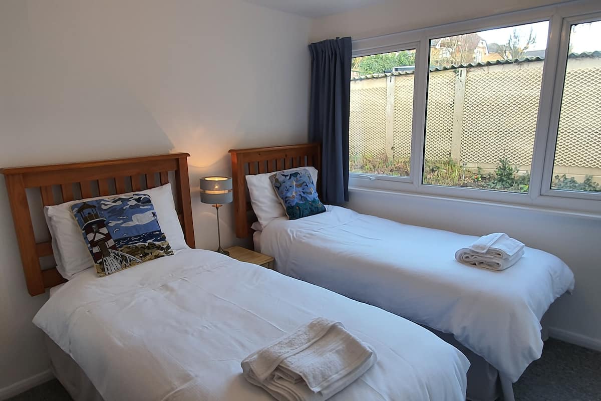 A hotel room with two single beds, each with a pillow and a folded towel. There is a nightstand with a lamp between the beds.