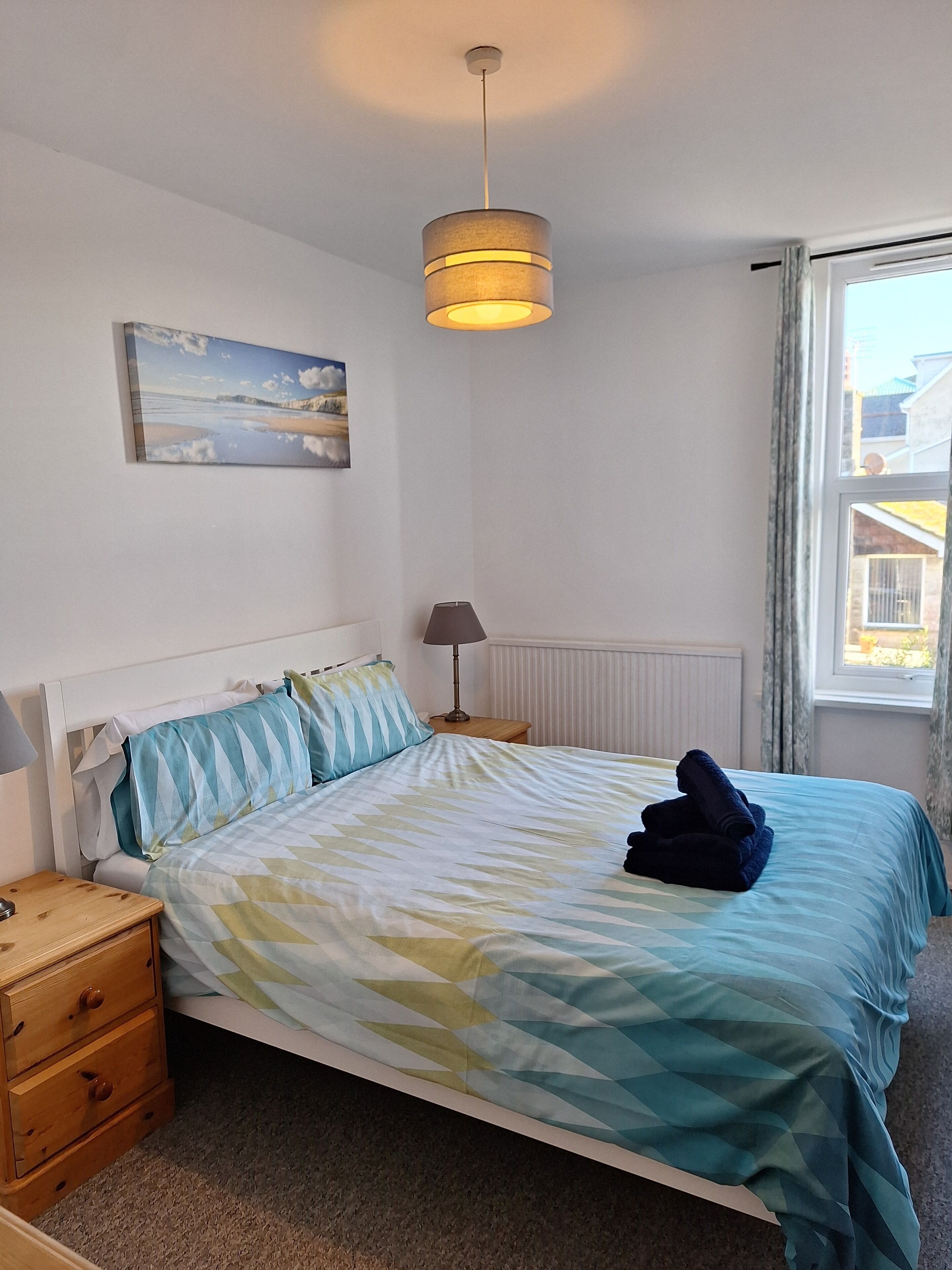 A bedroom with a bed, a nightstand, a lamp, a painting, and a window.