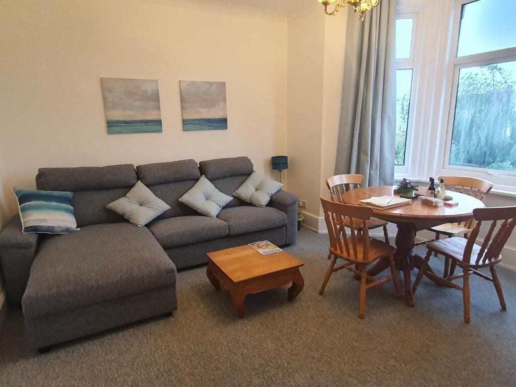 Ein Wohnzimmer mit einer Couch, einem Couchtisch und einem Esstisch mit Stühlen. An der Wand hängen zwei Gemälde und ein Fenster mit Vorhängen.