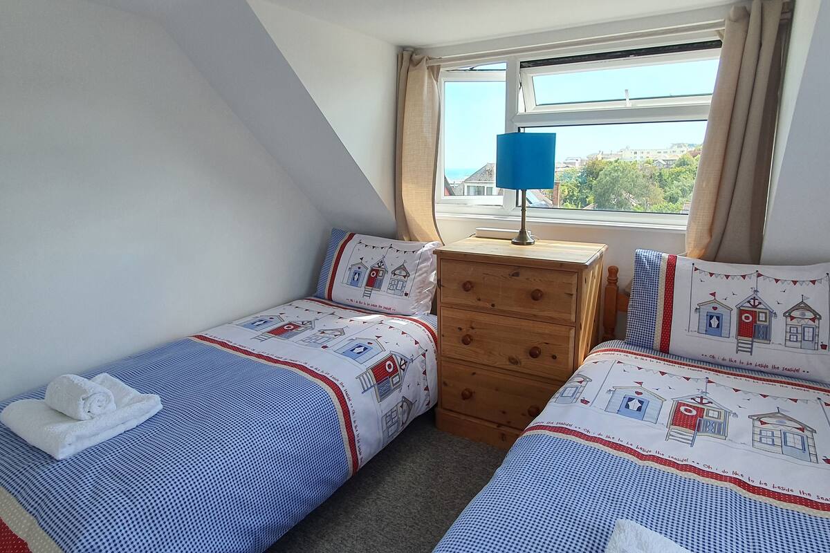 A bedroom with two single beds, a wooden dresser, a nightstand with a lamp, and a window with curtains.