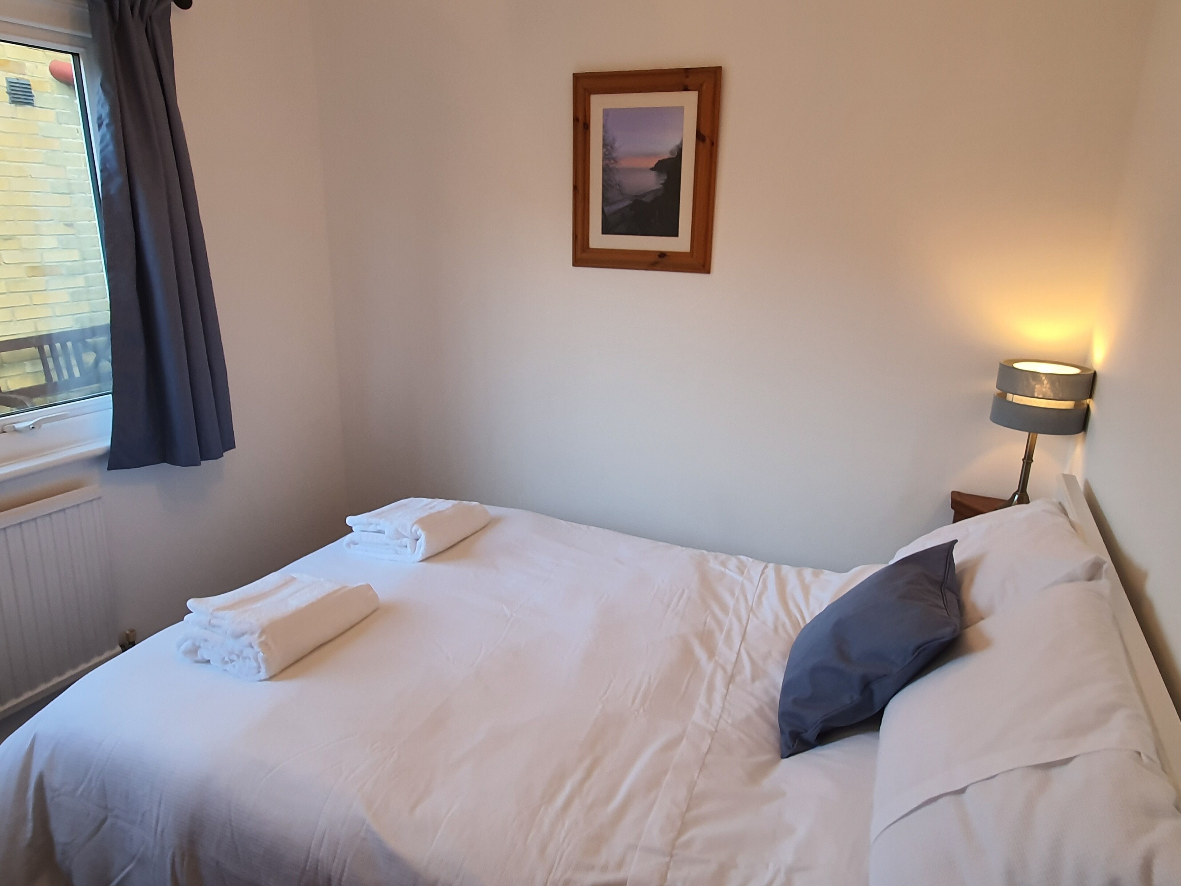 A neatly made bed with white linens and a folded towel, a bedside lamp, a framed picture on the wall, and a window with curtains.