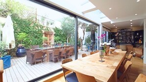 Un comedor moderno con una amplia mesa de madera y sillas, un muro de cristal que ofrece vistas a un patio exterior con mobiliario y plantas.