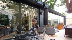 Un gimnasio en casa con equipo de ejercicio, incluyendo una bicicleta estática, una máquina de remo y una pesa rusa, en una terraza de madera con pared de vidrio que da a un jardín.