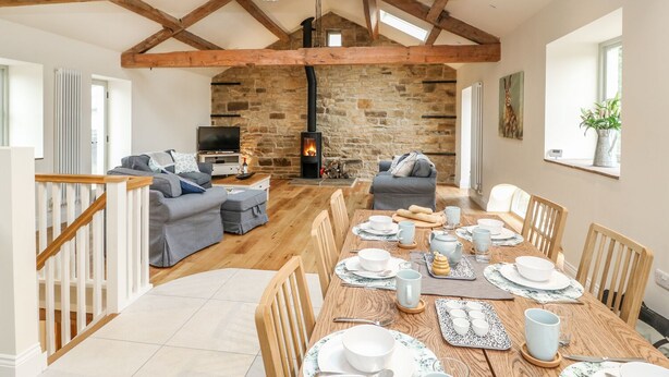 A spacious living room with a stone fireplace, wooden beams, a dining area set for four, and a cozy seating arrangement. - Morgan's Barn (Huddersfield)