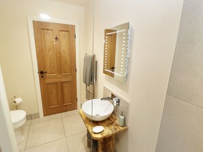 Un baño con puerta de madera, un lavabo blanco sobre un mueble de madera, un espejo con luces y una toalla colgada en un perchero.