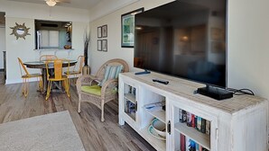 A living room with a large flat-screen TV, a dining table with chairs, a bookshelf, and a decorative mirror. - Retreat 615- Great Views, Nicely furnished, Pool, Easy Beach Access (Pawleys Island)