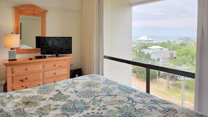 A hotel room with a bed, a TV, a dresser, and a large window with a view of buildings and greenery. - Retreat 615- Great Views, Nicely furnished, Pool, Easy Beach Access (Pawleys Island)