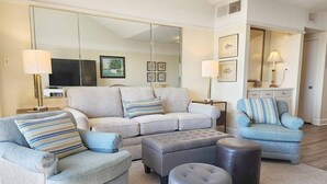 A modern living room with a sectional sofa, two armchairs, a grey ottoman, and a large mirror. - Retreat 615- Great Views, Nicely furnished, Pool, Easy Beach Access (Pawleys Island)